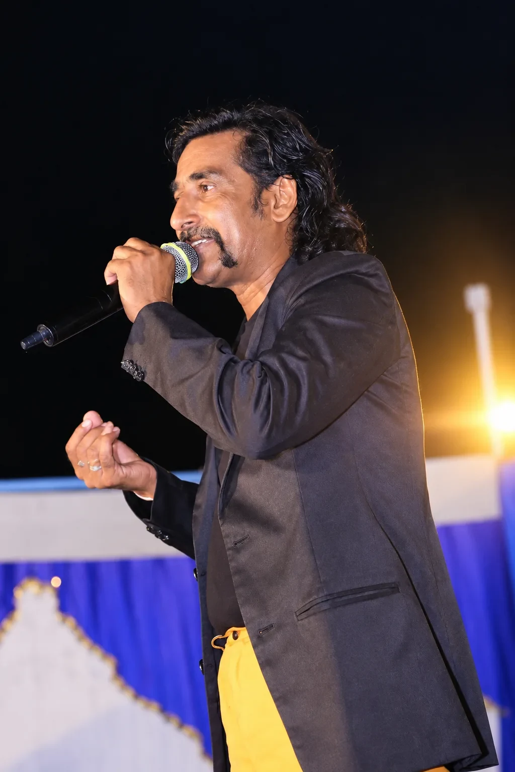 Jadugar Rajtilak speaking into the microphone in his black blazer and yellow shirt — poster frame from the official reel