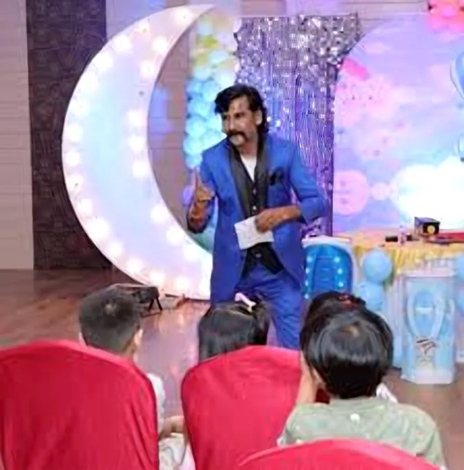 Jadugar Rajtilak performing live in his royal blue suit — children in red chairs watching under a glowing crescent-moon stage set